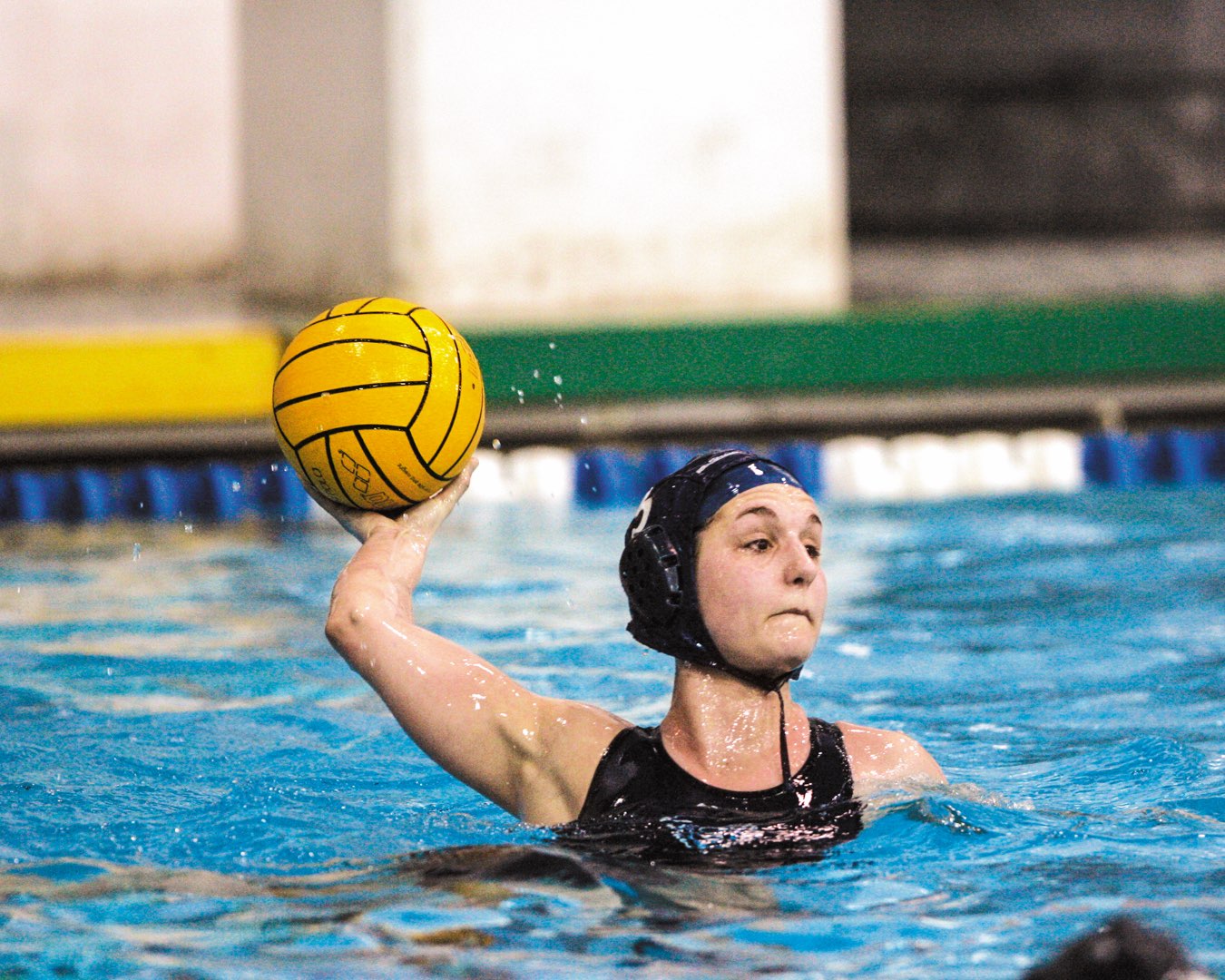 Villanova Women’s Water Polo player in her swim in the pool about to launch the bright yellow water polo ball