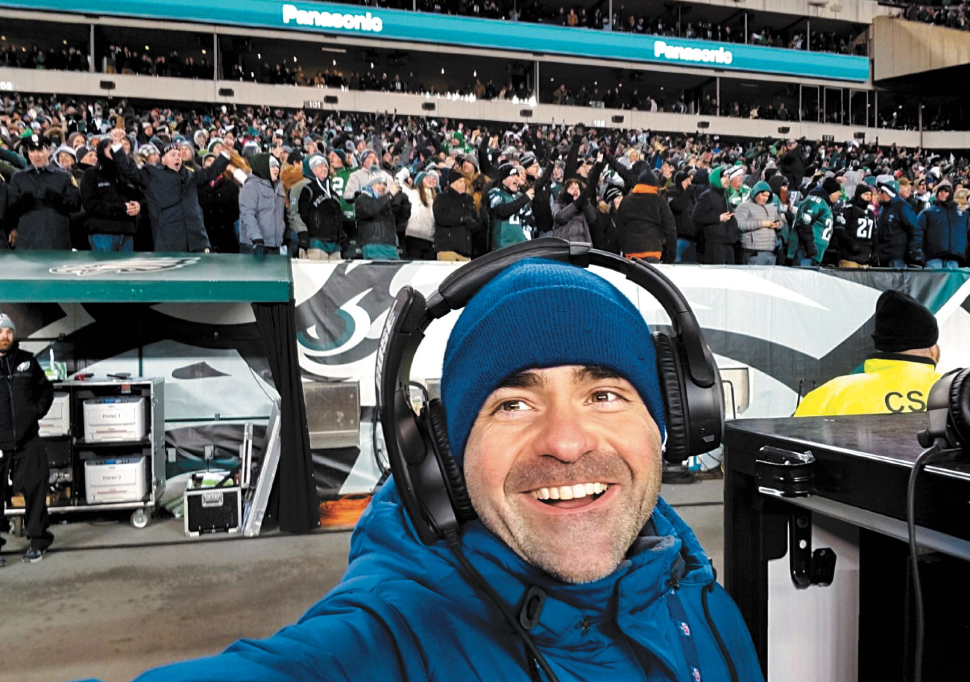 A selfie of CPS alumnus Ron Tedesco smiling in headphones on the sidelines with packed stands of Eagles fans behind him