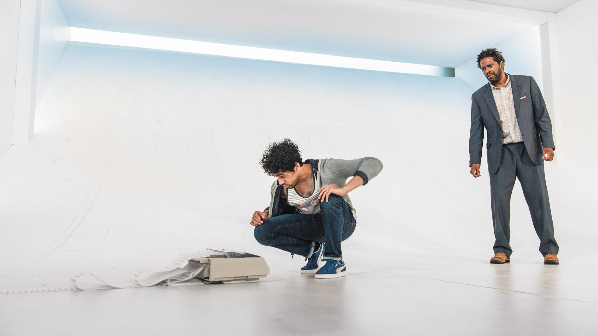 One young male crouched over a long list printing and a man in a suit standing against a stark white background