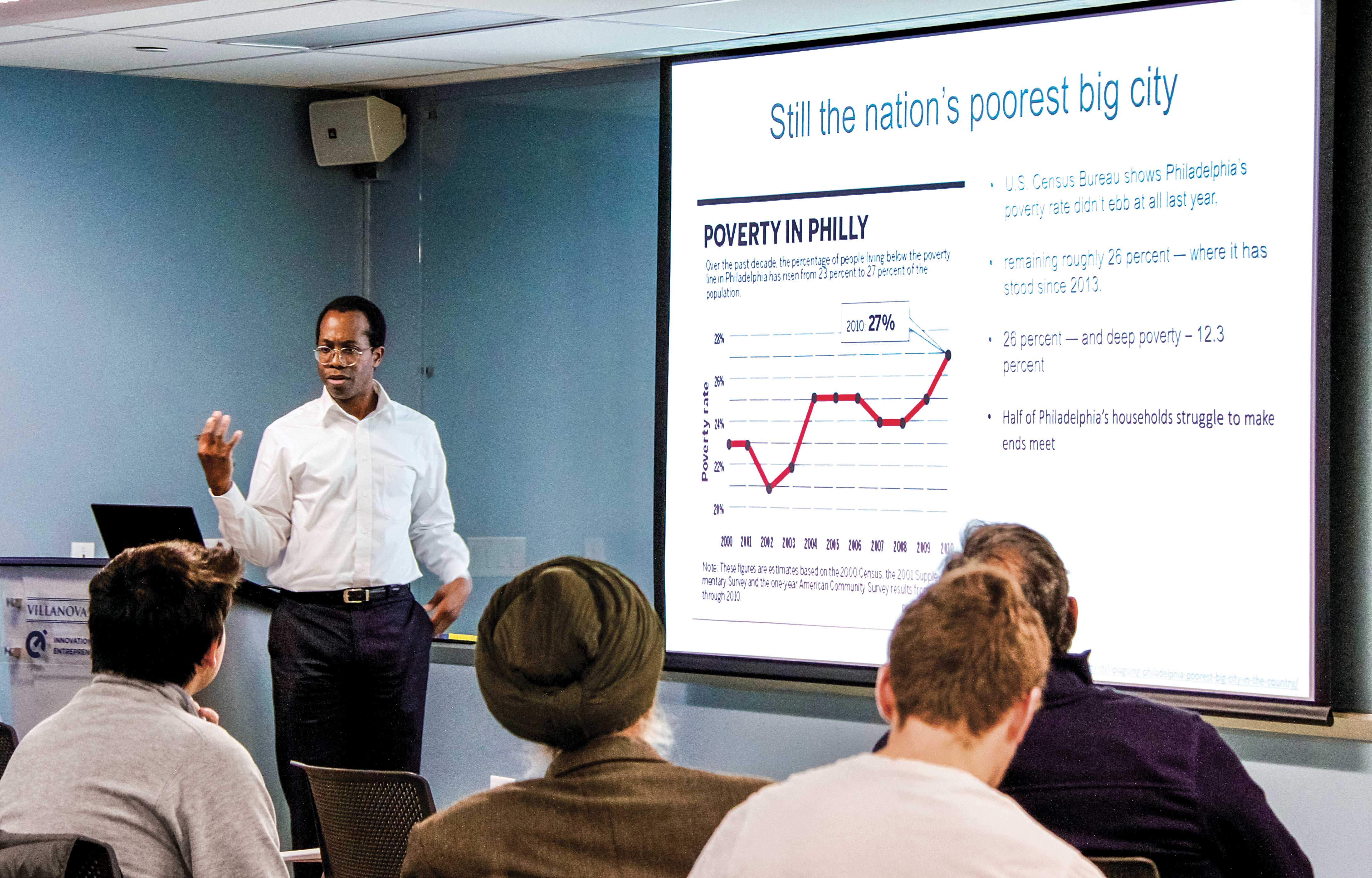 Alum and adjunct professor Hasshi Sudler talking to a class with a chart of poverty in Philly on the projector screen