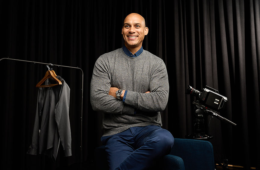 Joe Tribble, backstage in front of a black curtain, sits on the arm of a chair with his arms and legs crossed 